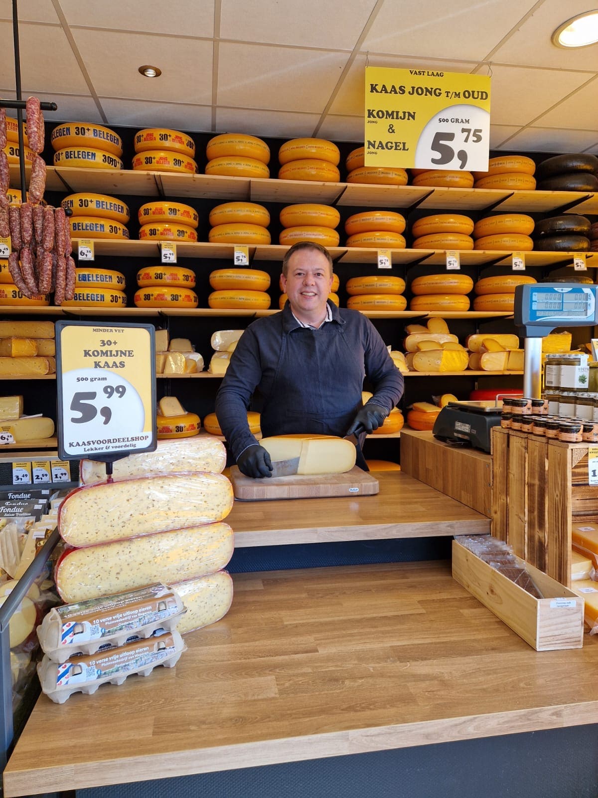 Ondernemer van Kaasvoordeelshop Burgum snijdt verse kaas achter de toonbank, met de kaaswand op de achtergrond